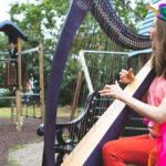A woman in bright colours sits on a bench in a children's play park playing the harp. To her left, two more people are playing in the play park.