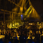 Yellow spotlights illuminate an orchestra, different sections on different platforms within the audience in a vast hall.