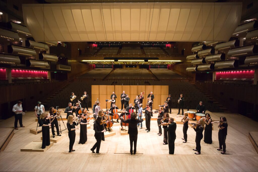 A chamber orchestra stand on the stage of an empty concert hall, playing their instruments