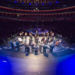 A birds eye view image of an orchestra with their backs to the camera. In front of them stands a conductor, with his face to the camera