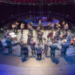 A birds eye view image of an orchestra with their backs to the camera. In front of them stands a conductor, with his face to the camera