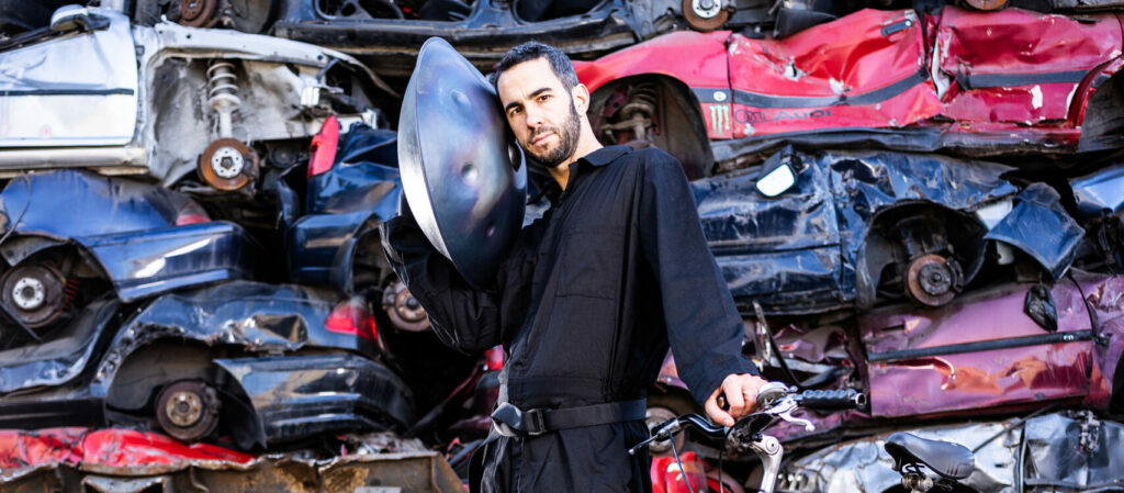A man stands holding a handpan up to his right ear, and holding a bicycle by its handlebars, in front of a huge pile of crushed cars of different colours.
