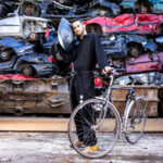 A man stands holding a handpan up to his right ear, and holding a bicycle by its handlebars, in front of a huge pile of crushed cars of different colours.