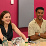 A woman and a man sit at a table smiling, in front of a pink wall