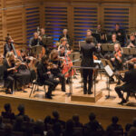 A full orchestra are performing on a stage, with audience members sat in darkness watching them.