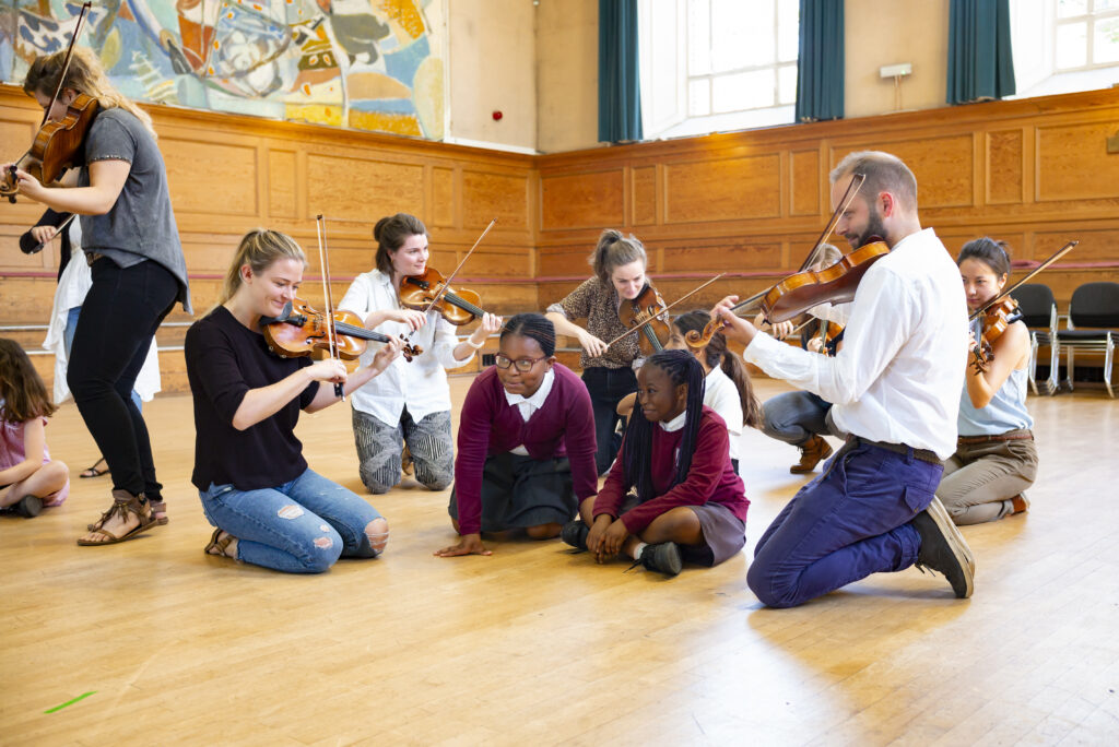 Two students in school uniform sit in amongst five violinists.