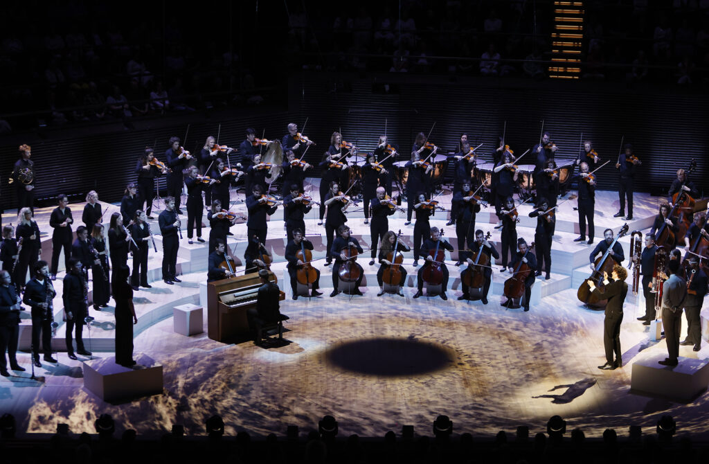 An orchestra stand in two lines on a stage. In front of them sit a line of cellists. A conductor stands to one side, with his arms up conducting. On the left hand side of the stage is a piano. The stage is lit up with projections, including a big black spot in the middle of the stage.