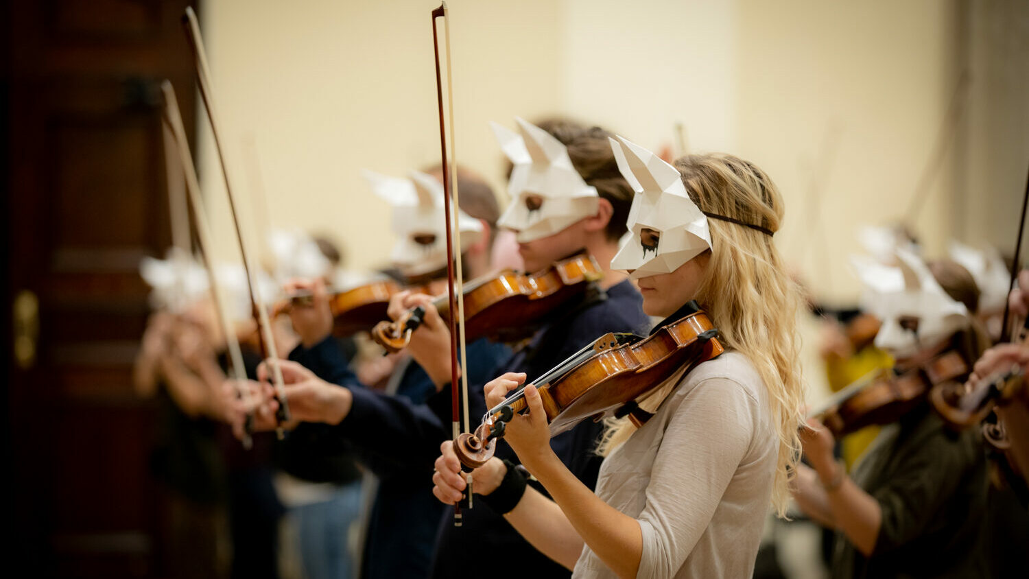 Violinists stand in a diagonal line, with white masks on. Their bows are held up, with their violins on their left shoulder.