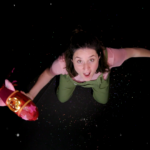 A woman stands looking up at the camera above her head, a look of awe on her face. Her arms are out to the sides. In her right arm she holds a pink rocket.