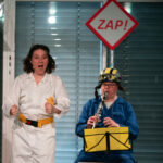 A woman in a white boilersuit wearing a yellow seatbelt stands with her hands in a driving position, looking excited. To her right sits a man in a blue boilersuit and a yellow racing car hat, playing a clarinet. in front of him is a music stand with a yellow piece of paper on it. Behind him is a big red and white sign that reads 'Zap!'