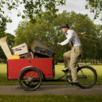 A man sits on a bicycle with a trailer full of musical items in front of it. He is carrying a trombone on his back. He is in a park.